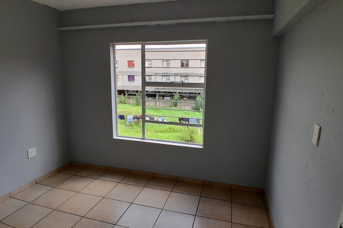 A vacant bedroom featuring neutral grey walls, tiled flooring, and a large window with security bars overlooking the exterior grounds.