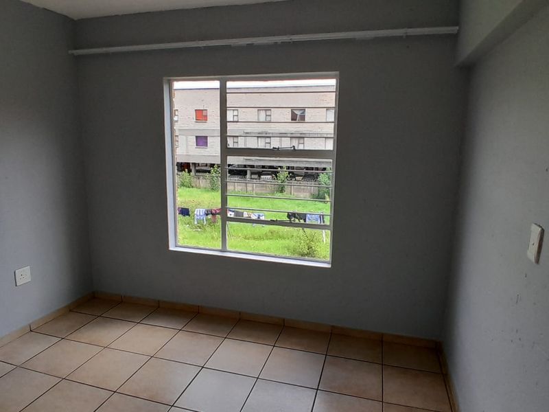 A vacant bedroom featuring neutral grey walls, tiled flooring, and a large window with security bars overlooking the exterior grounds.