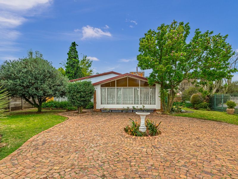 A well-maintained rear exterior view featuring a brick-paved courtyard, mature garden landscaping, and a house with a gabled roof and large glass windows.