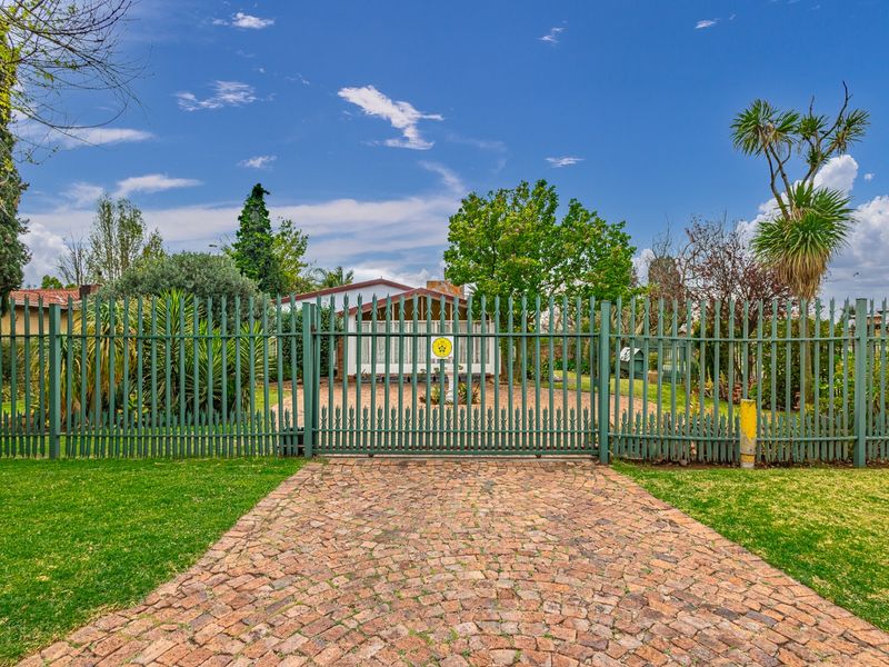 Front view of the property featuring a paved driveway, secure metal fencing, and a well-maintained garden area.