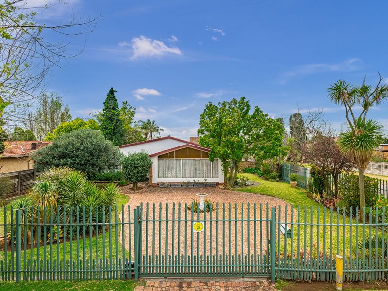 Front view of a residential property featuring a paved driveway, established garden, and secure metal perimeter fencing.