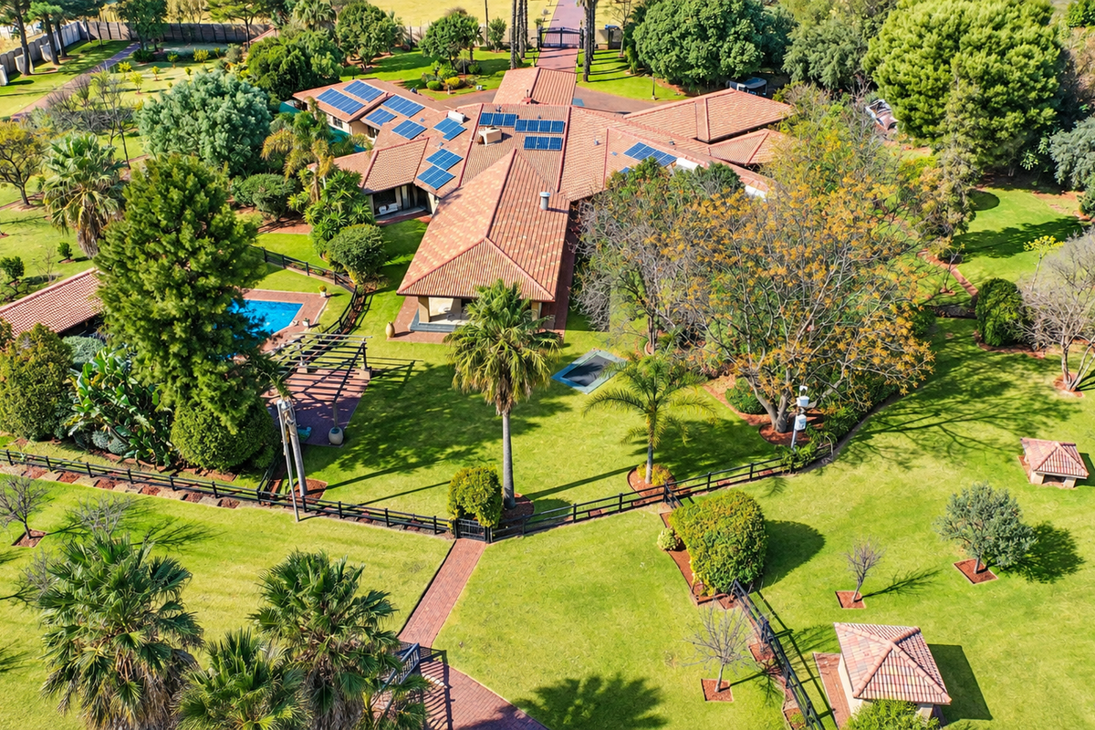 Aerial view of a spacious small holding featuring a large residence with solar panels, a swimming pool, extensive landscaped gardens, and paved pathways.