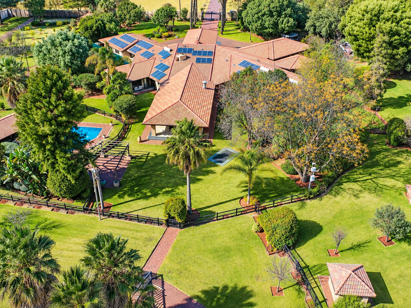 Aerial view of a spacious small holding featuring a large residence with solar panels, a swimming pool, extensive landscaped gardens, and paved pathways.