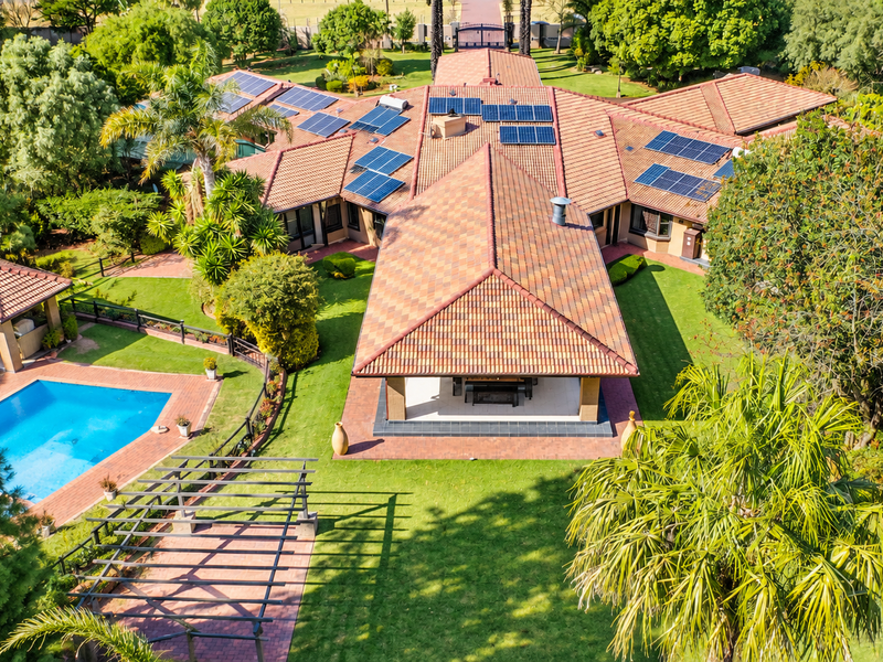 Aerial view of a large property featuring a tiled roof with solar panels, a swimming pool, a covered patio, and extensive landscaped gardens.