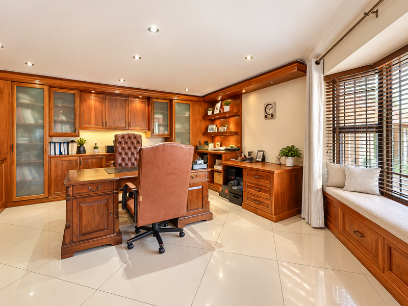 A well-appointed home office featuring extensive custom-built wooden cabinetry, a comfortable bay window seat, and polished tile flooring with recessed lighting.