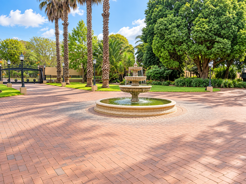 A grand entrance featuring a paved driveway, a central tiered water fountain, mature palm trees, and secure wrought iron gates.