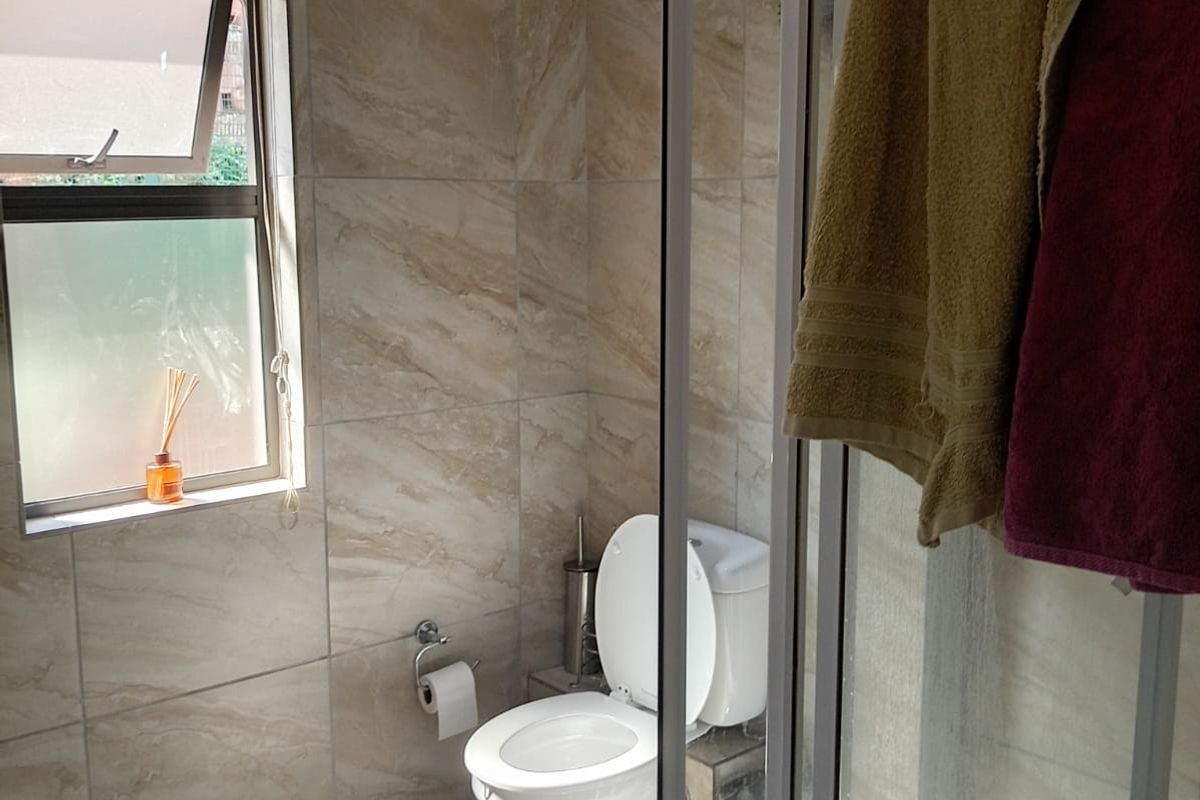 A functional bathroom featuring floor-to-ceiling tiled walls, a glass shower enclosure, and a toilet, with natural light provided by a side-facing casement window.