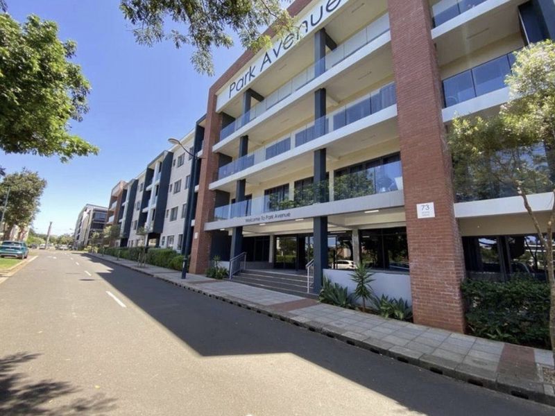 Street-level view of the Park Avenue apartment complex, featuring a modern brick facade, glass balconies, and a paved pedestrian entrance.