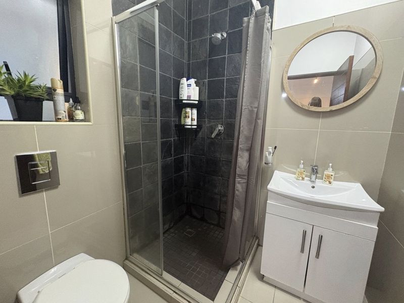 A compact modern bathroom featuring a tiled shower enclosure, a white vanity cabinet with a washbasin, and neutral wall tiling.