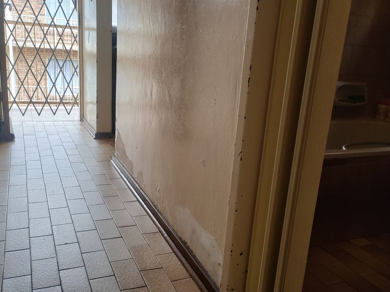 A narrow hallway with tiled flooring and visible wall surface deterioration, leading towards a security gate and an adjacent bathroom.