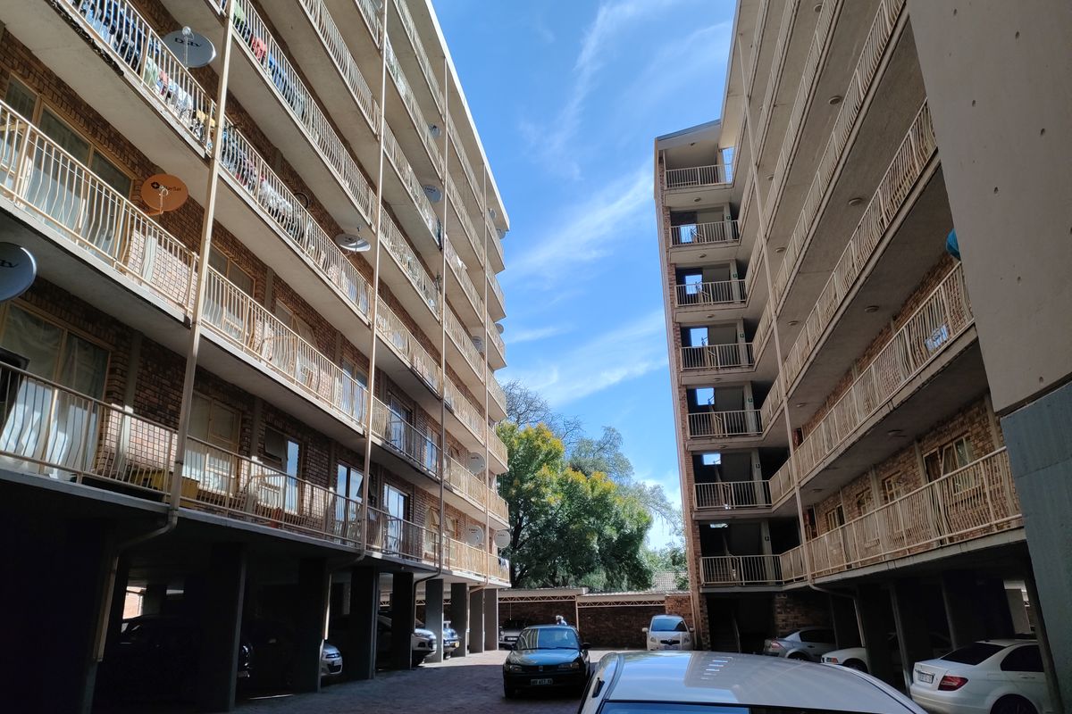 Exterior view of a multi-story apartment complex featuring balconies with metal railings and a paved ground-level parking area.