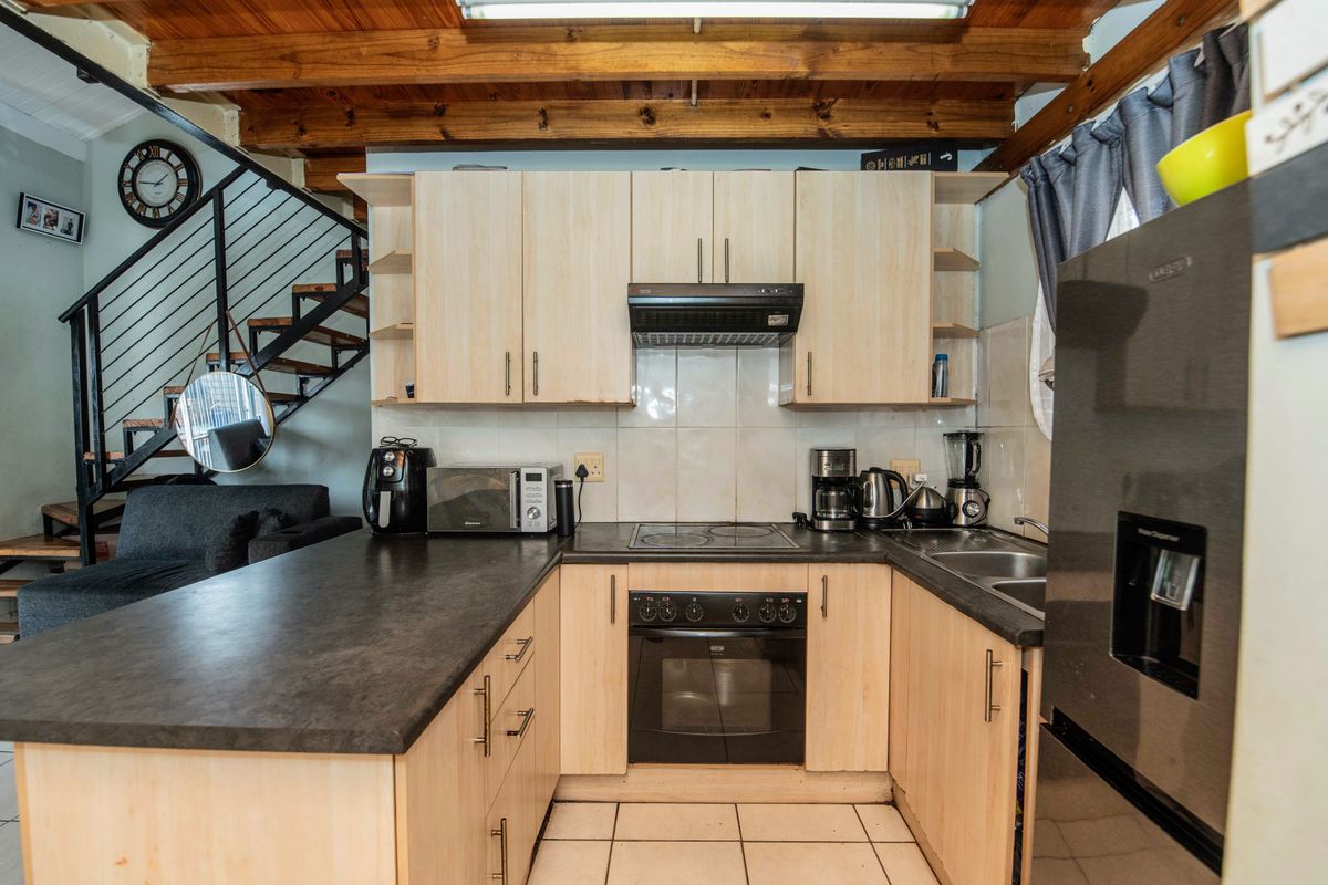 A functional kitchen featuring light wood cabinetry, dark laminate countertops, built-in oven and hob, and exposed timber ceiling beams, adjacent to an open staircase.