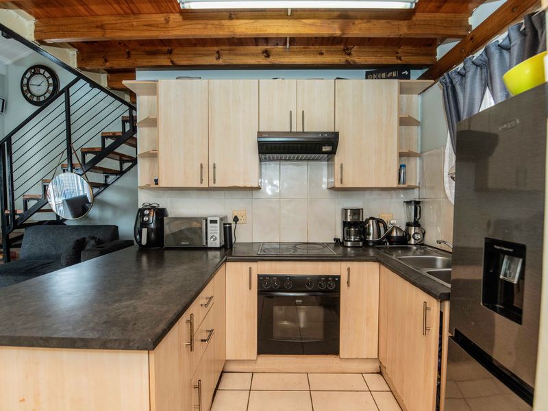 A functional kitchen featuring light wood cabinetry, dark laminate countertops, built-in oven and hob, and exposed timber ceiling beams, adjacent to an open staircase.