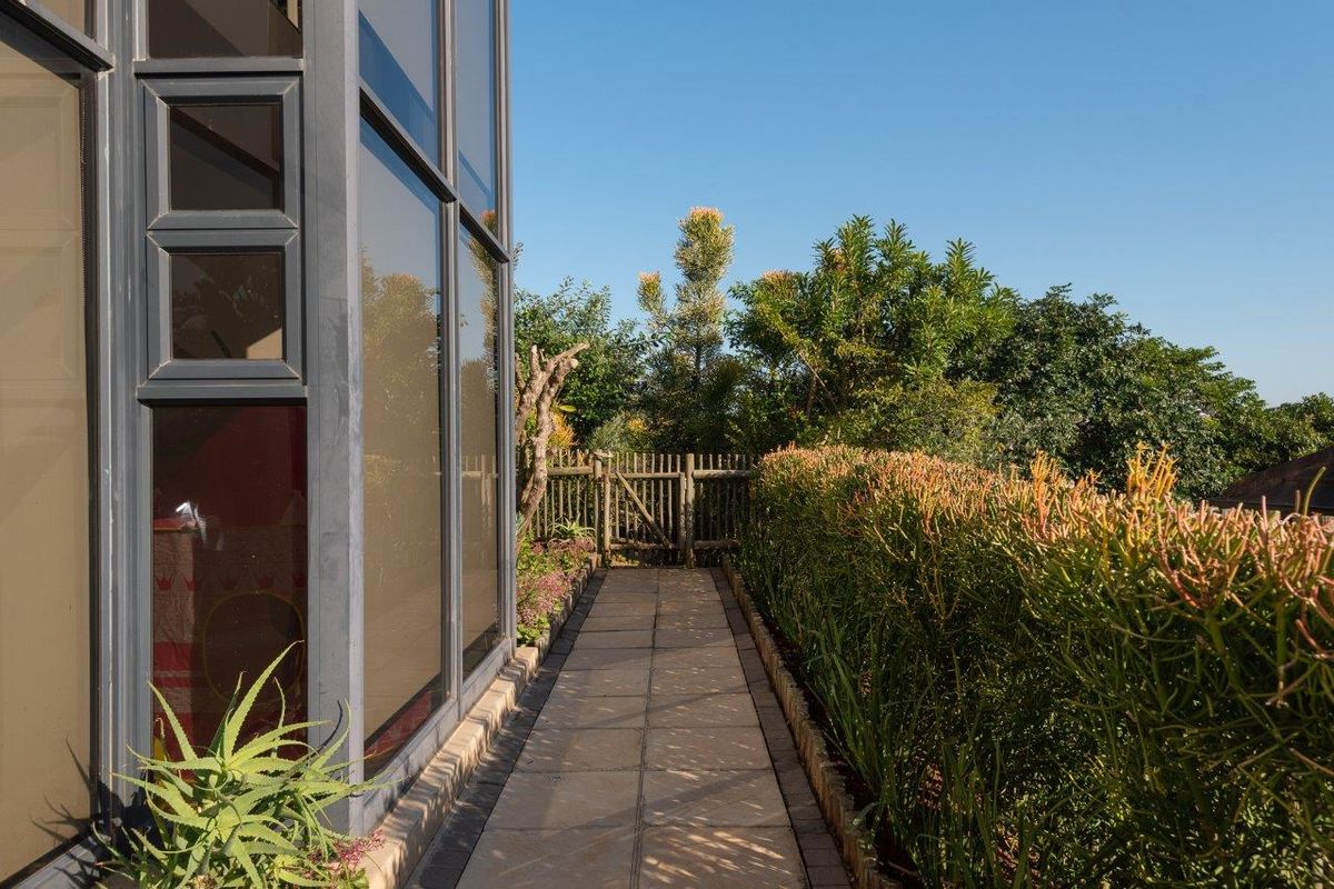 A well-maintained paved walkway leads through a landscaped garden area alongside the property, featuring mature hedges and large glass windows.