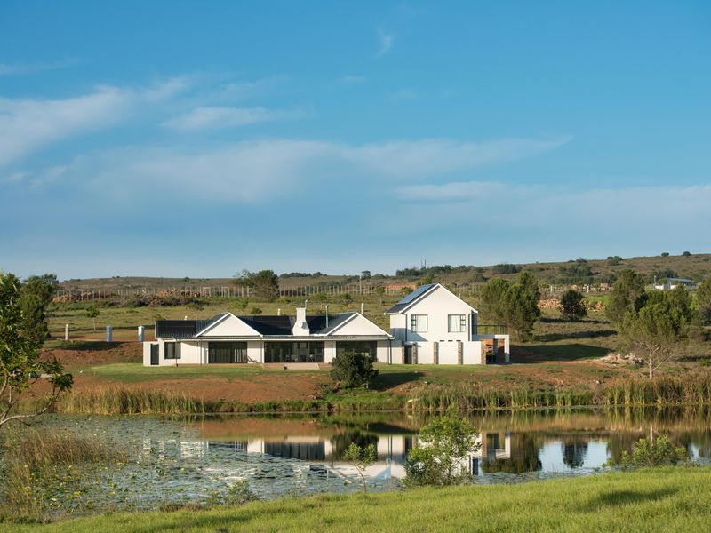 A scenic view of the rear elevation of a modern residential property featuring a pitched roof, solar panels, and a patio overlooking a pond.