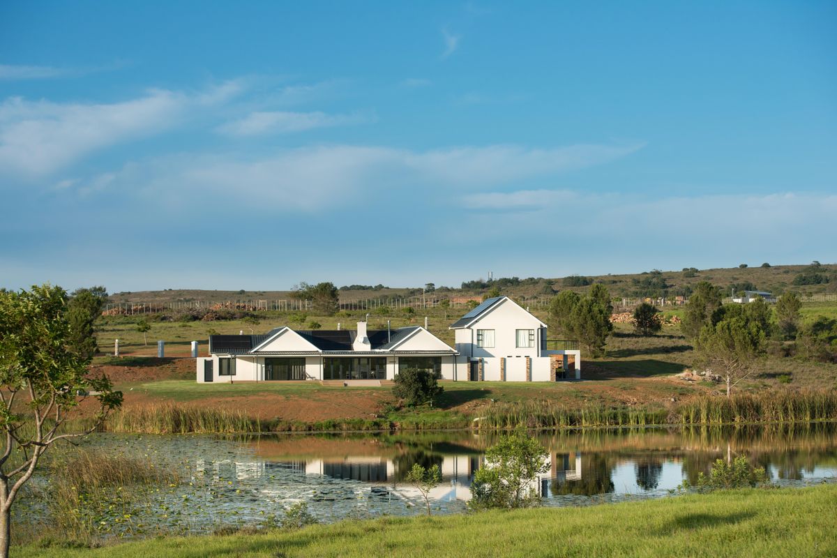 A scenic view of the rear elevation of a modern residential property featuring a pitched roof, solar panels, and a patio overlooking a pond.