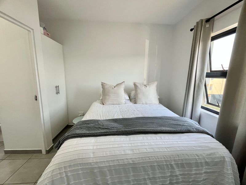 A compact bedroom featuring tiled flooring, a built-in wardrobe, and a window with neutral curtains.
