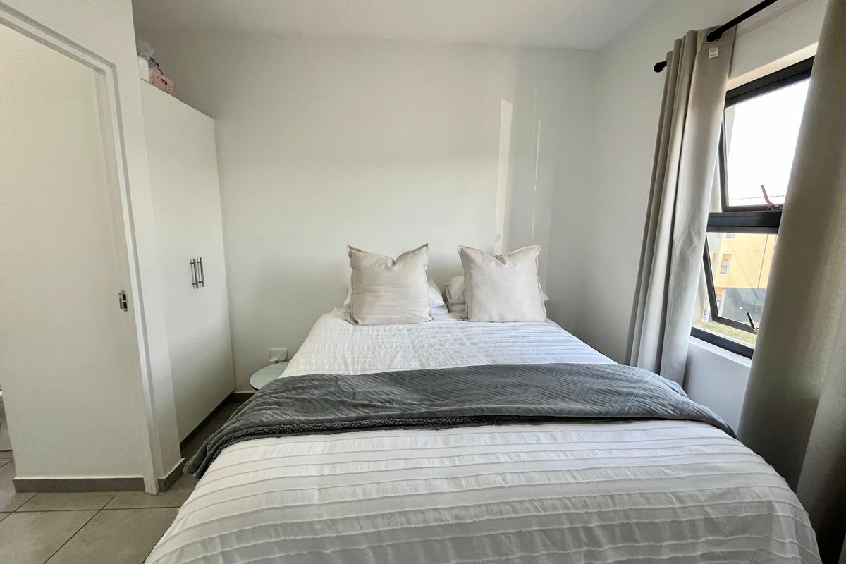A compact bedroom featuring tiled flooring, a built-in wardrobe, and a window with neutral curtains.