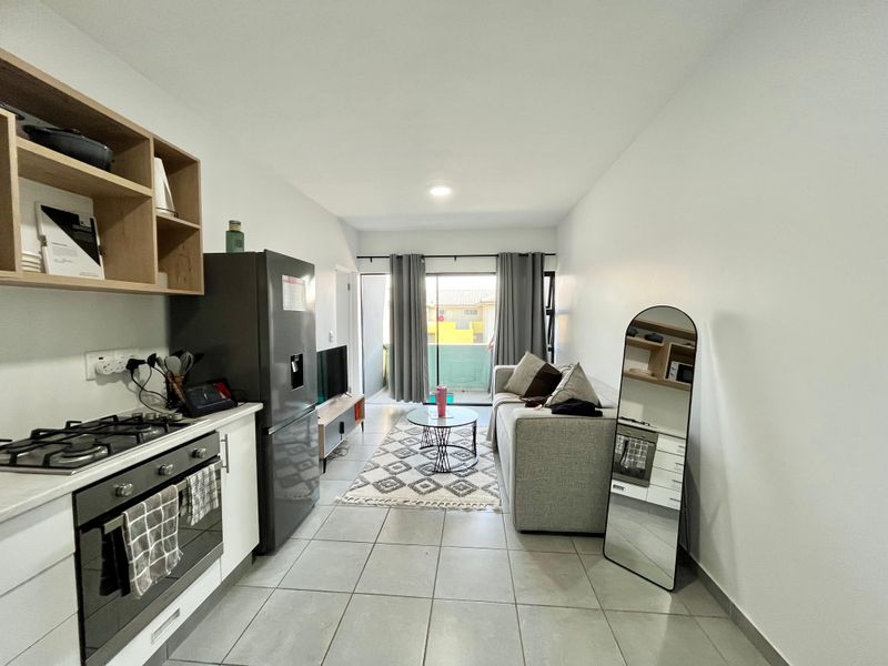 Modern open-plan living area featuring a compact kitchen with built-in oven and gas hob, flowing into a lounge space with access to a balcony.