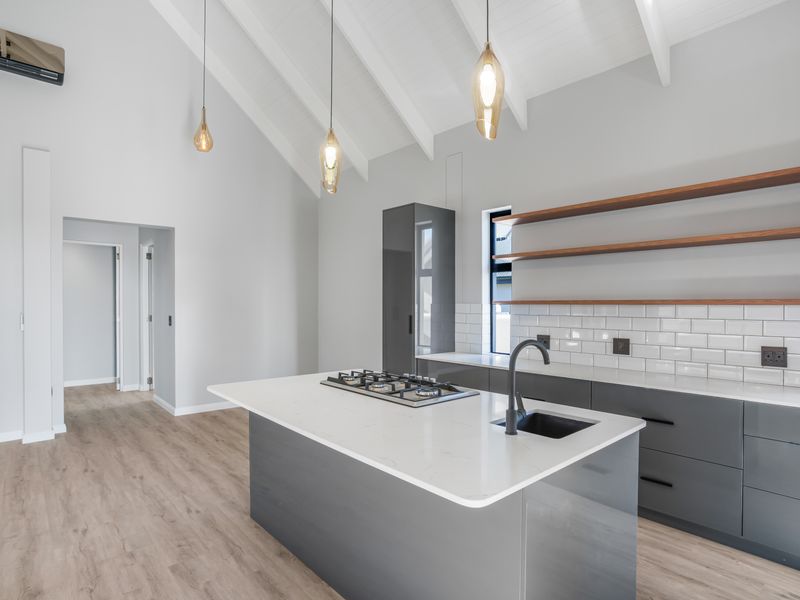 Modern kitchen featuring a central island with gas cooktop, sleek grey cabinetry, subway tile backsplash, floating timber shelves, and a striking vaulted ceiling.