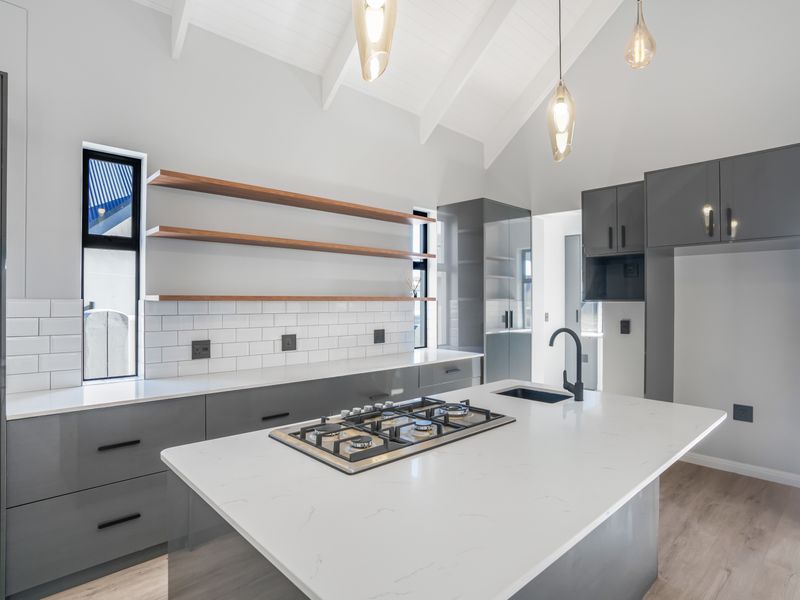 Modern kitchen featuring a central island with gas cooktop, sleek grey cabinetry, subway tile backsplash, floating timber shelves, and high vaulted ceilings.