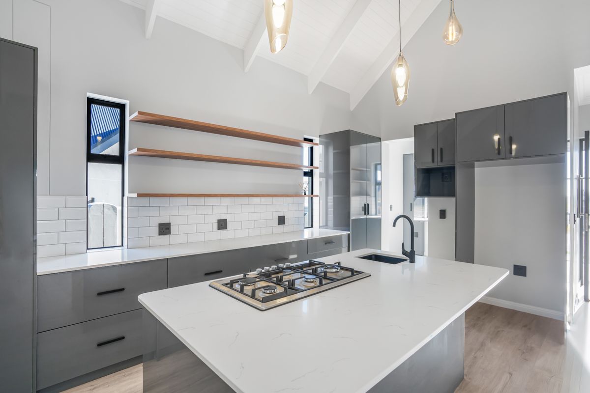 Modern kitchen featuring a central island with gas cooktop, sleek grey cabinetry, subway tile backsplash, floating timber shelves, and high vaulted ceilings.