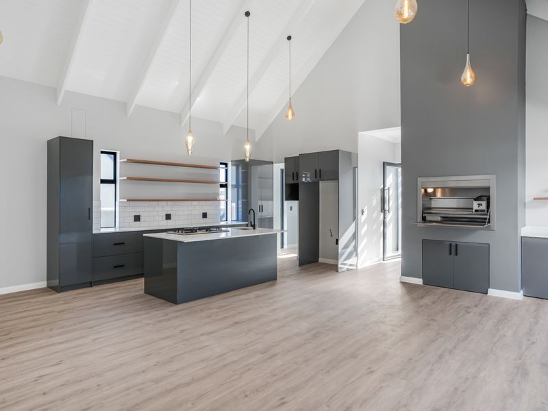 Modern open-plan kitchen and living area featuring a vaulted ceiling, sleek grey cabinetry, stone countertops, a central island, and a built-in indoor braai.