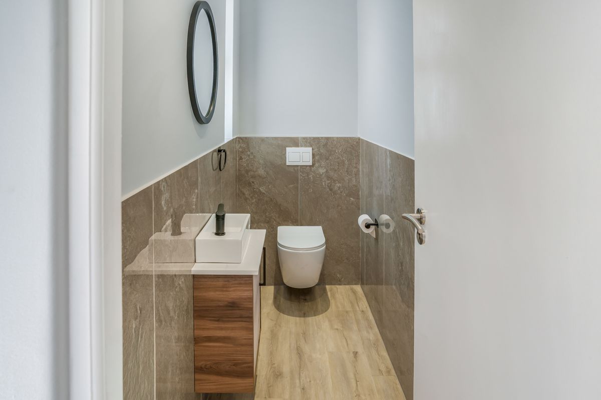 Modern guest bathroom featuring a wall-mounted toilet, wood-look vanity cabinet with vessel sink, and neutral stone-look wall tiling.