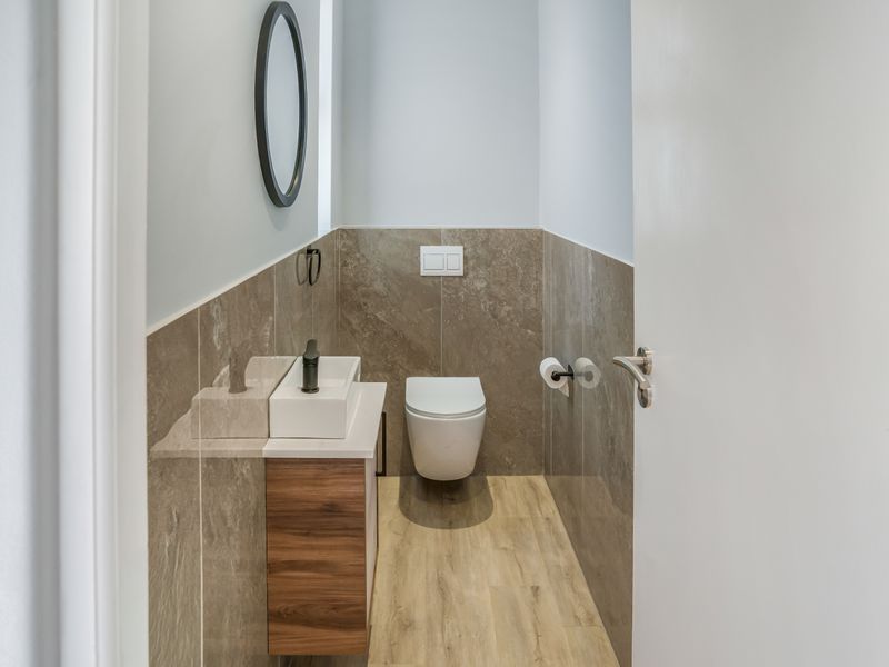 Modern guest bathroom featuring a wall-mounted toilet, wood-look vanity cabinet with vessel sink, and neutral stone-look wall tiling.