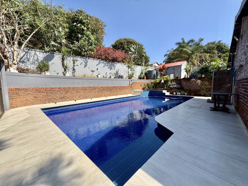 A well-maintained outdoor swimming pool area featuring a tiled surround, brick retaining walls, and established garden landscaping.