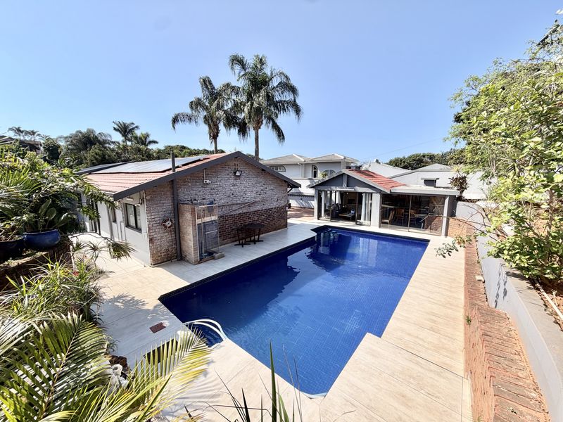 A well-maintained outdoor pool area featuring a tiled deck, brick-clad building exterior, and solar panels on the roof, set within a landscaped garden.