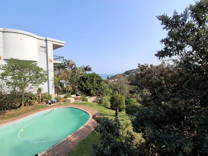 An elevated view of a swimming pool with brick coping, set within a landscaped garden alongside a multi-story residential building with distant coastal views.