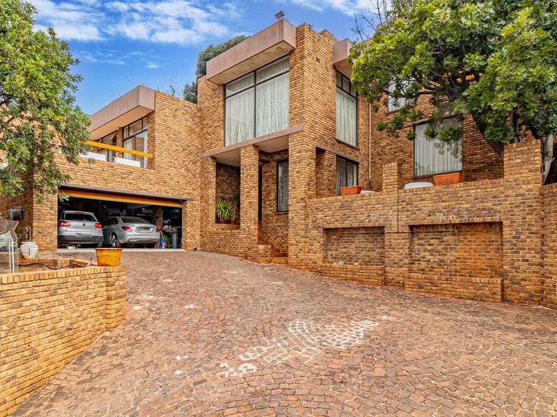 A distinctive multi-level face brick residence featuring a large cobbled driveway, integrated double garage, and expansive windows set against a landscaped hillside.