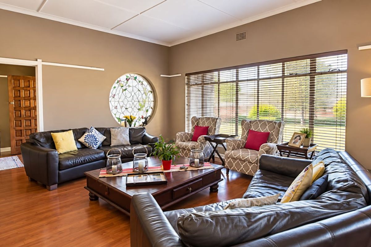 Spacious living room featuring warm laminate flooring, a unique circular feature window, and large windows with venetian blinds allowing for ample natural light.