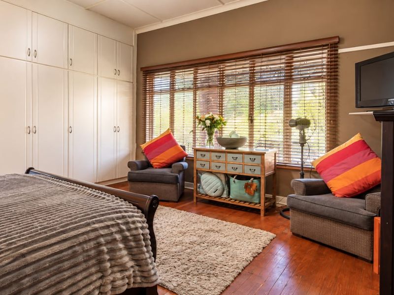 Spacious bedroom featuring extensive built-in cabinetry, warm wooden flooring, and large windows with venetian blinds, offering direct access to an en-suite bathroom.