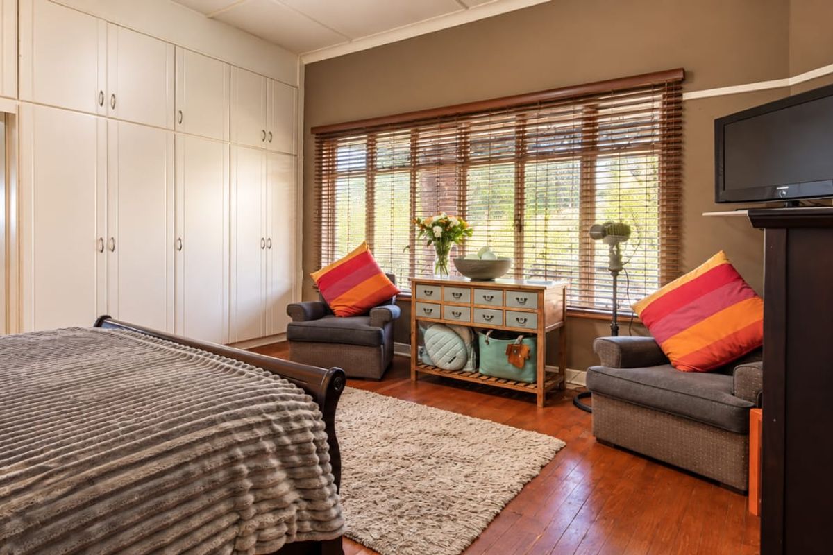 Spacious bedroom featuring extensive built-in cabinetry, warm wooden flooring, and large windows with venetian blinds, offering direct access to an en-suite bathroom.