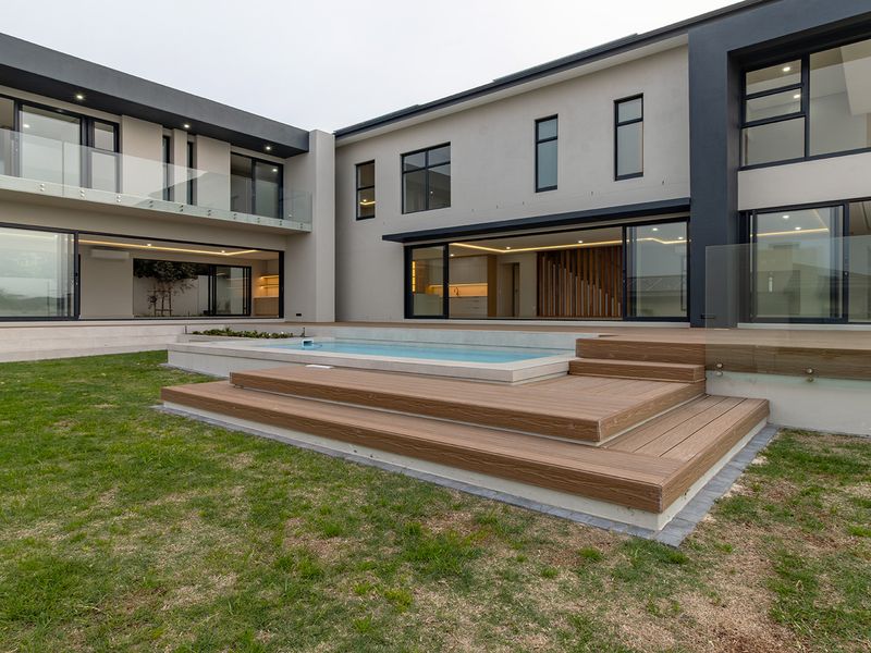 Modern rear exterior featuring a swimming pool, timber decking, glass balustrades, and expansive sliding doors connecting the indoor and outdoor living spaces.