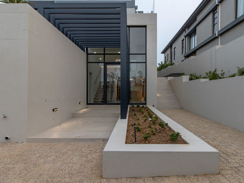 Modern front entrance featuring a sleek pergola, glass door, concrete planter box, and paved driveway with integrated exterior lighting.