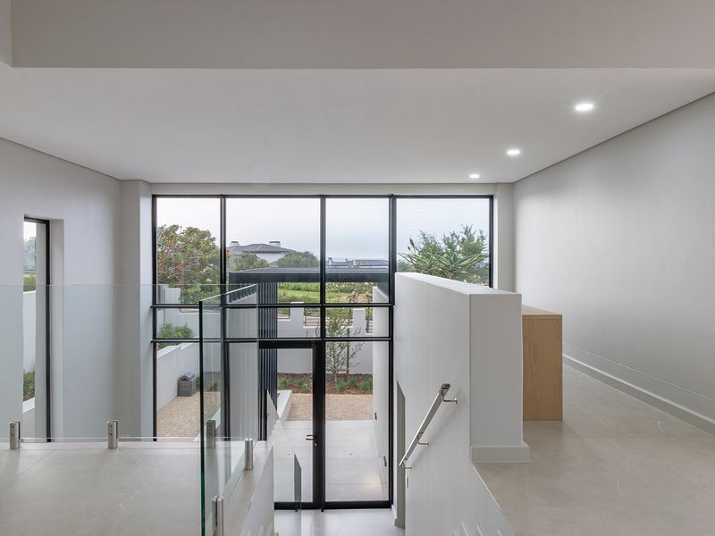 A bright, modern hallway featuring large format floor tiles, a sleek glass balustrade, and expansive windows overlooking the exterior.