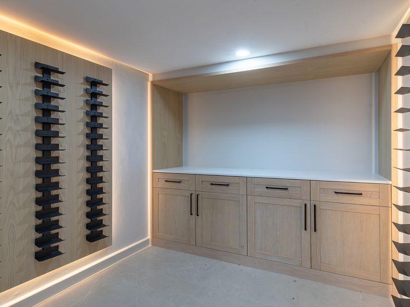 A modern, custom-designed wine cellar featuring light wood cabinetry, stone countertops, and integrated metal wine racking with ambient LED lighting.