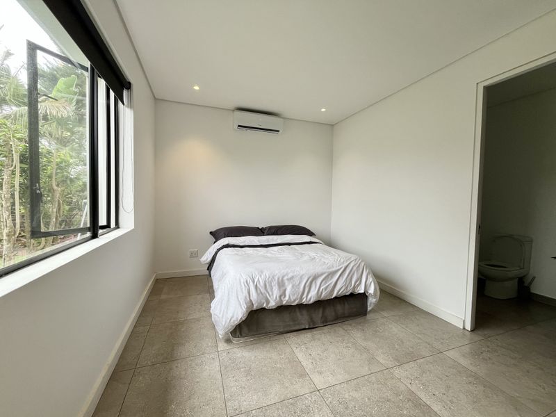 A clean, modern bedroom featuring neutral tiled flooring, recessed lighting, a wall-mounted air conditioning unit, and direct access to an en-suite bathroom.