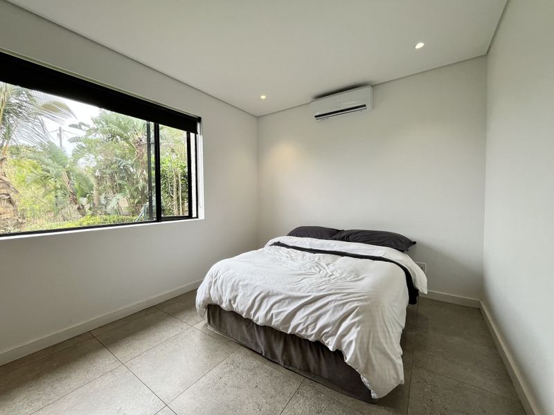 A bright and modern bedroom featuring neutral tiled flooring, recessed lighting, and a wall-mounted split-system air conditioner with a large window overlooking greenery.