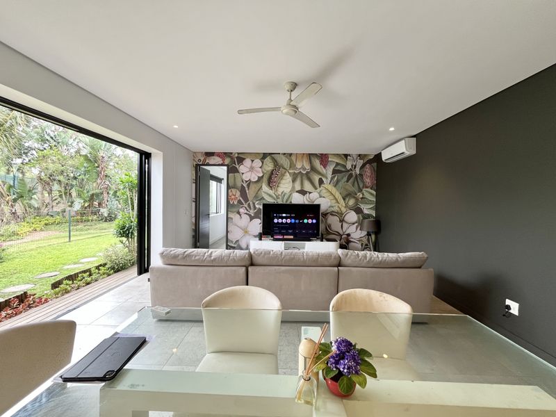 Modern open-plan living area featuring large sliding doors opening to a garden, a ceiling fan, air conditioning, and a decorative feature wall.