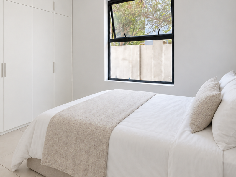 A bright and modern bedroom featuring neutral tones, built-in floor-to-ceiling cupboards, and large aluminium-framed windows providing natural light.