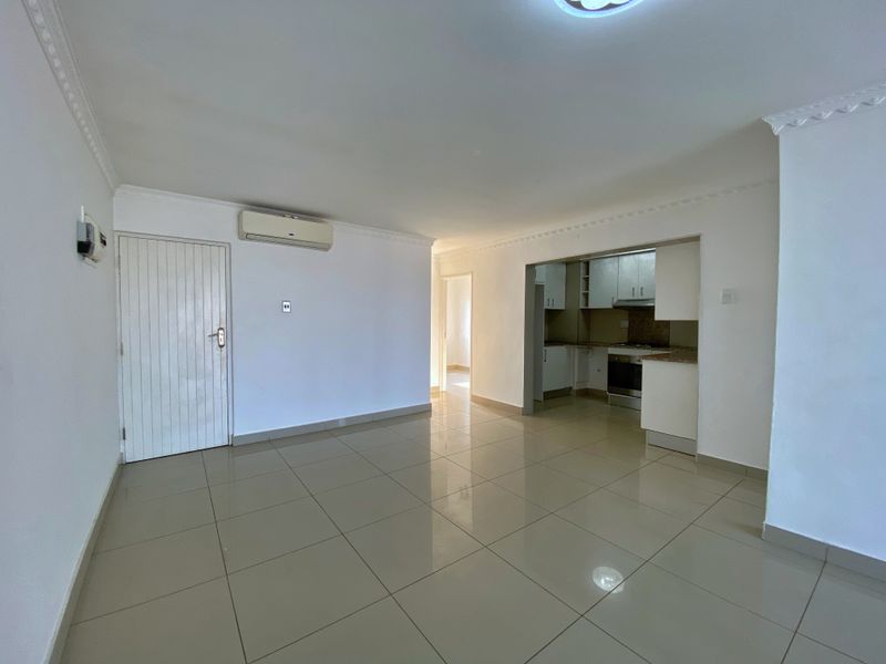 An open-plan living area featuring tiled flooring, a wall-mounted air conditioner, and an adjoining kitchen with built-in cabinetry and appliances.