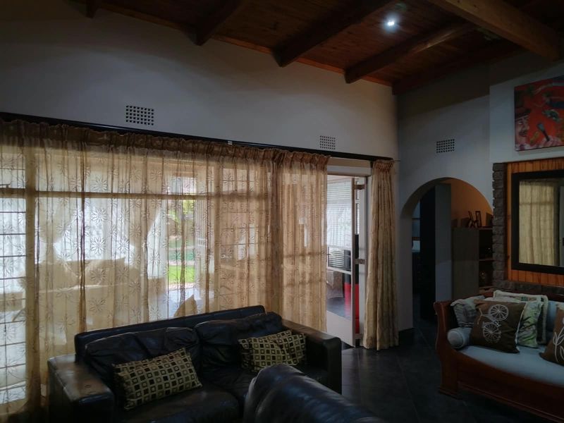 Spacious living room featuring exposed timber beam ceilings, tiled flooring, and an arched entryway leading to adjacent areas with access to the patio.
