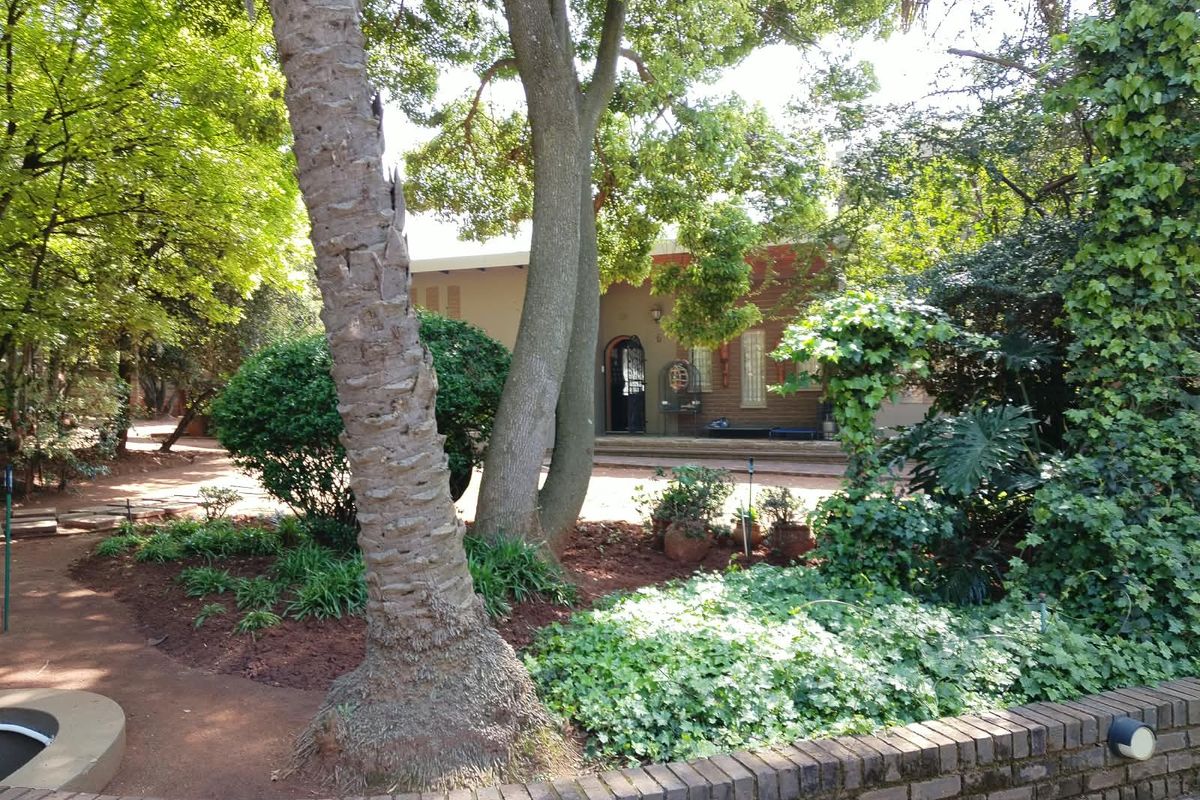A view of the rear garden featuring mature trees, landscaped beds, a brick retaining wall, and a paved patio area leading to the house entrance.
