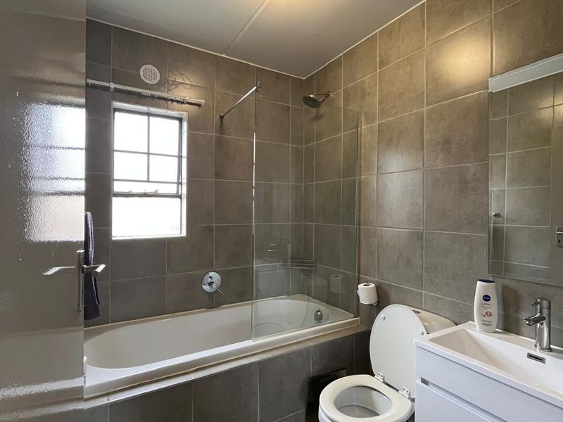 A modern bathroom featuring a bathtub with a glass shower screen, a wall-mounted vanity with a basin, and neutral floor-to-ceiling wall tiles.