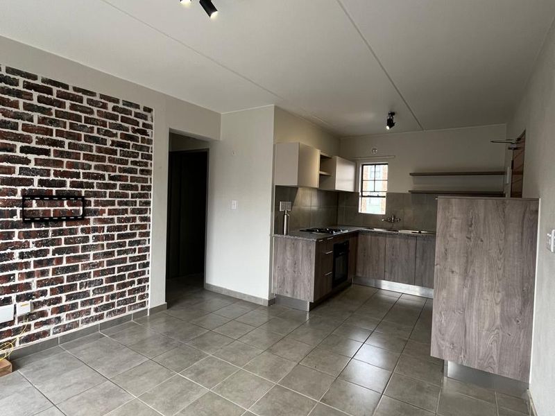 Open-plan living and kitchen area featuring a rustic exposed brick wall, neutral floor tiles, and modern wood-finish cabinetry with integrated appliances.