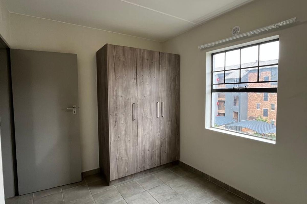 A clean, empty bedroom featuring neutral tiled flooring, a built-in wooden wardrobe, and a window providing natural light.